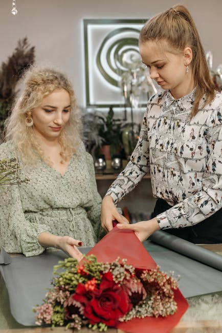 A florist, possibly a professional from Wandsworthflowers.org.uk, is arranging a floral gift on a table behind a bouquet featuring vibrant red roses, pink and white filler flowers, and greenery, wrapp