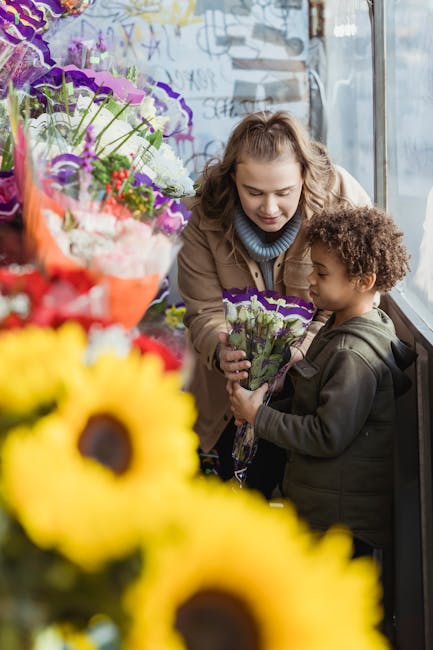 A young woman with wavy blonde hair and a light blue turtleneck sweater is handing a small bouquet of purple and white flowers, wrapped in purple paper, to a child with curly brown hair wearing a dark
