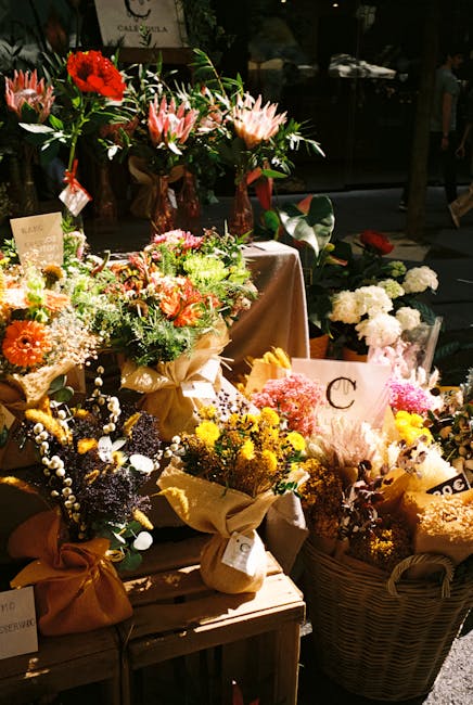 A vibrant display of mixed floral arrangements and bouquets arranged on a wooden table and in woven baskets, featuring a variety of fresh flowers including roses, carnations, gerbera daisies, and lili